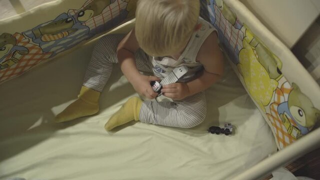 A fair-haired preschool boy playing with toy cars while sitting in a crib. Top view of a kid doing leisure activity. Close-up. Home bedroom interior.