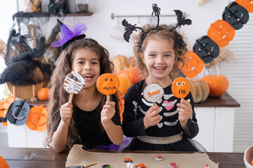 two diverse kids girl in costume of witch, having fun in kitchen, eating cookies, celebrating...
