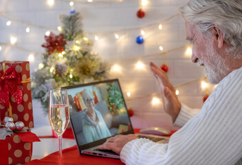 Naklejka na ściany i meble Portrait of old senior man using laptop to video call with distant people. He celebrates the Christmas event with a glass of sparkling wine and smiling. Gift box and Christmas tree on the table