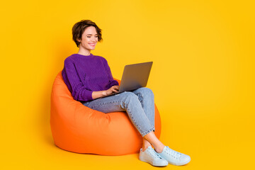 Full length photo portrait of woman working on laptop sitting in orange beanbag chair isolated on vivid yellow colored background