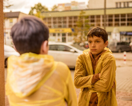 Photo of teenage boy in yellow raincoat among city streets looks into reflection of glass windows, sadness loneliness autumn depression