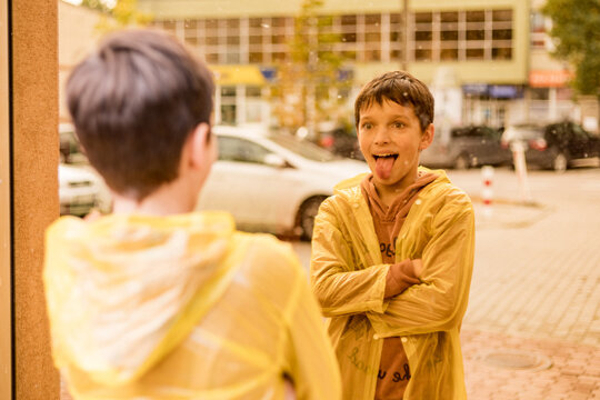 Photo of teenage boy in yellow raincoat among city streets looks into reflection of glass windows, sadness loneliness autumn depression, Sticking Out Tongue