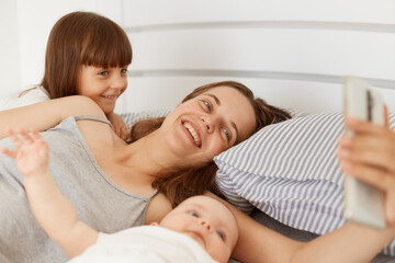 Indoor shot of happy smiling female wearing gray sleeveless t shirt lying in bed with her children and holding smartphone in hands, having video call with husband.