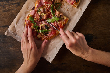 Women's hands cut with knife rectangular Roman pizza with prosciutto ham, tomatoes, mozzarella,...