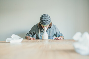 Ill man with long hair and hand made wool hat using a steam inhaler on wooden table during flu season - obrazy, fototapety, plakaty