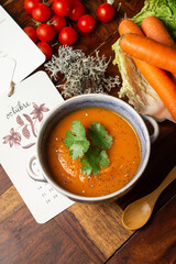 Aerial view of bowl with carrot cream with coriander leaves, on wooden table with carrots, tomatoes and calendar in Spanish, vertical