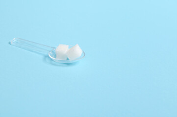Fototapeta na wymiar Pure refined white sugar cubes on a transparent measuring spoon isolated over colored blue background with copy space for medical advertising. World diabetes day awareness concept, 14 November.