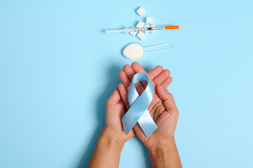 Top view of hands holding blue ribbon, symbol of World Diabetes Awareness Day on blue background with insulin syringe and pure refined white sugar cubes. Concept of dangers of eating white sugar
