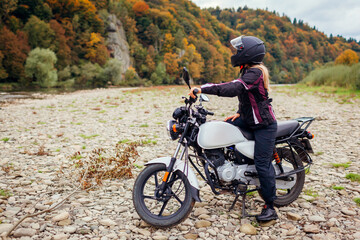 Woman biker travel by motorbike in fall. Motorcyclist enjoys autumn landscape in mountains having rest by forest