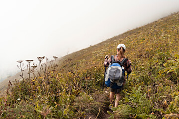 rear view of young woman in plaid shirt with big backpack with trekking poles walking along mountain trail hiking in fog, healthy active lifestyle