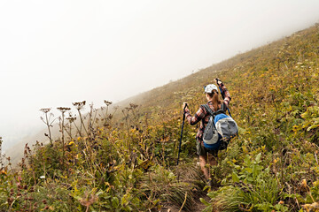 Naklejka na ściany i meble rear view of young woman in plaid shirt with big backpack with trekking poles walking along mountain trail hiking in fog, healthy active lifestyle