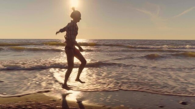 A slender woman runs and jumps on the waves of sea water on a sandy beach. on the surf. Splashes of water and foam on the background of the orange sun at sunset. Caucasian beauty is happy and joyful.