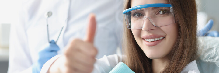 Young woman in safety glasses sitting in dental chair and showing thumbs up