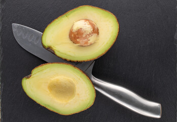 Two halves of an organic ripe avocado with a metal knife on a slate stone, close-up, top view.