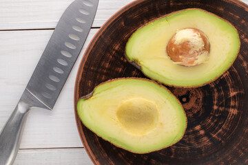 Two halves of organic ripe avocado on a ceramic dish with a metal knife on a wooden table,...