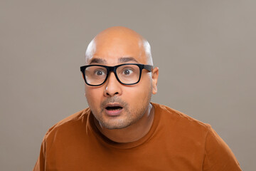 Portrait of a bald man wearing eyeglasses gaping with amazed expression against plain background.