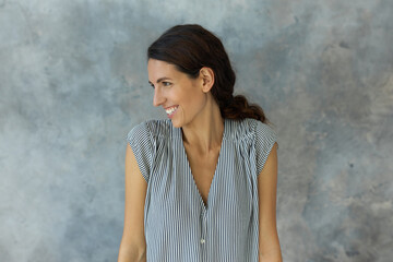 Positive young attractive Caucasian brunette woman standing in front of camera with head turned to right side, laughing, keeping hands down straight, dressed in casual shirt-sleeved lovely dress