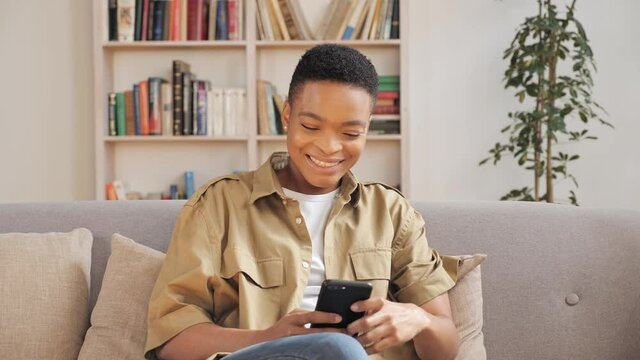 Cheerful middle age african woman using smartphone while sitting on the couch. Black smiling woman using app on cellphone at home. Beautiful girl relaxing chatting messaging on mobile phone indoor.