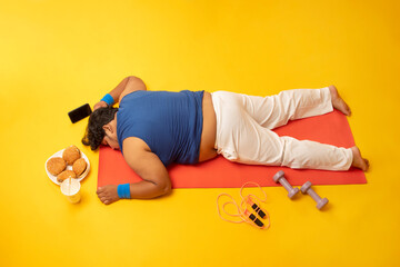 A fat man sleeping on yoga mat amidst dumbles,skipping rope,Burger,Drink and mobile phone.