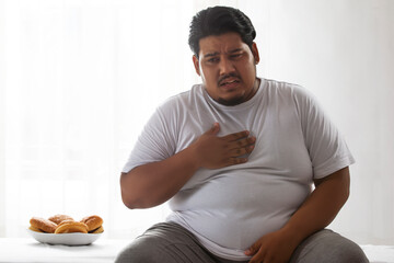  A fat man sitting,keeping hand on his chest due to pain with a plate of burger aside.