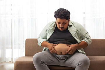  A fat man sitting on couch  holding and looking at extra belly fat.