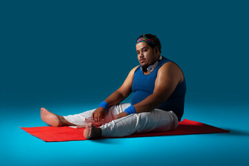 A fat man sitting on a yoga mat with headphones around neck exhausted after a workout.