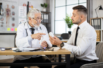 Professional male doctor sitting at table with caucasian businessman and giving advices how to take pills. Concept of medication and prescription.