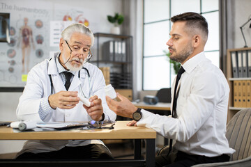 Naklejka na ściany i meble Mature therapist in white lab coat and eyewear consulting male patient in formal clothes about pills. Doctor prescribing medicine while sitting at desk with caucasian businessman.