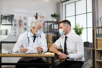 Competent aged doctor in face mask sitting at desk with caucasian businessman and holding sterile...