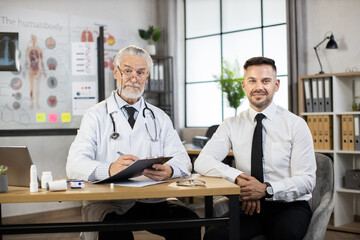 Naklejka na ściany i meble Portrait of caucasian man in business clothes sitting at desk with aged doctor that writing on clipboard. Therapist and patient looking at camera. Health examination concept.