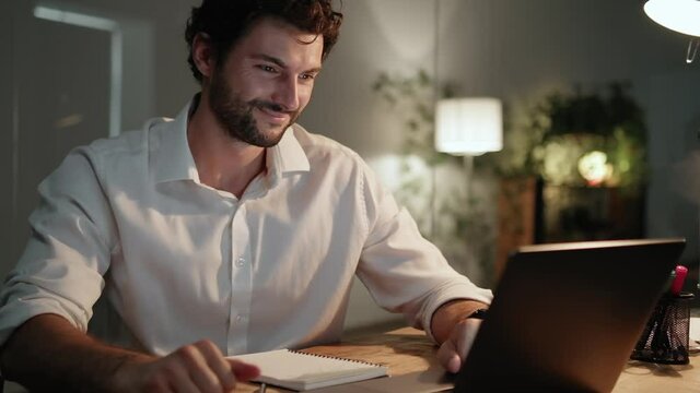A smiling man talking on the video-call on laptop in the office at night