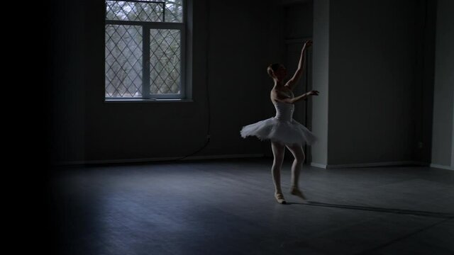 Wide shot slim ballerina in white dancing in darkness walking away leaving. Young beautiful Caucasian woman in tutu making steps on tiptoes. Choreography art and rehearsal concept