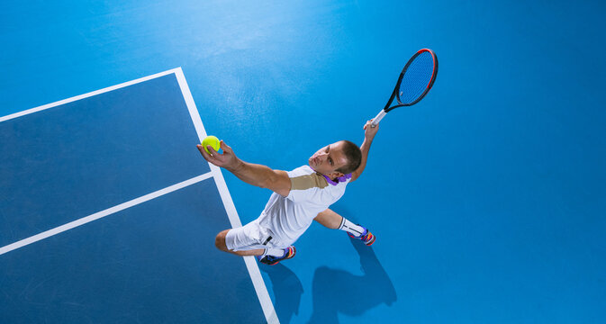 Full-length top view portrait of male professional tennis player serving a ball, training over gym background.
