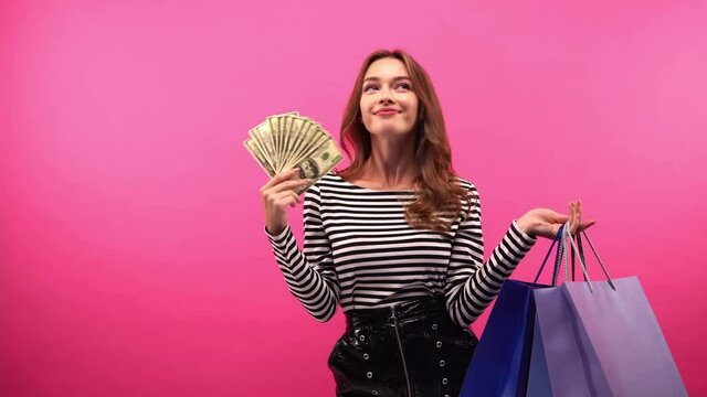 happy woman holding shopping bags and dollars isolated on pink