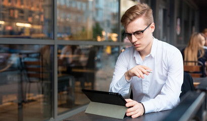 Young handsome blond guy sits with a tablet on the terrace of a cafe, works or studies in a public place