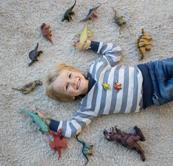cute boy lies on light carpet on his back. There are many dinosaur figures spread out around it. little scientist, children's hobby - prehistoric animals. interesting games for children. Fun childhood © Anna