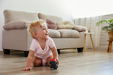 Portrait of smiling one year old child playing with a toy car on wooden floor of living room. Interior background. Adorable blonde little girl in pink crawlers at home. Close up, copy space for text.