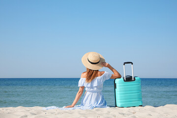 Woman with suitcase sitting on sandy beach near sea, back view