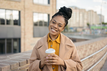 Beautiful smiling African American woman wearing stylish coat holding cup of coffee looking at camera on the street