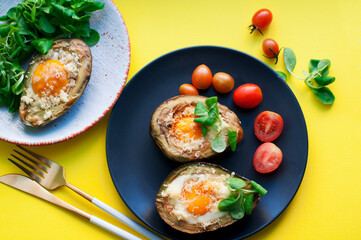 Baked avocado with egg, cheese, corn salad and tomatoes on black and white plates on yellow background. Breakfast with avocado. Gold knife and fork. Top view