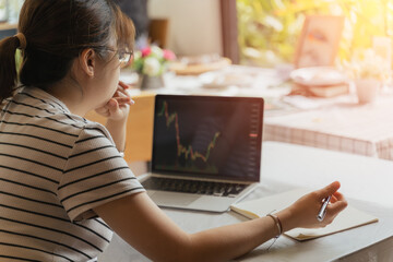 Young adult Asian trader, investor, businesswoman using computer laptop with graph chart in business money investing, stock market, Bitcoin Cryptocurrency trading at home. Financial investment concept