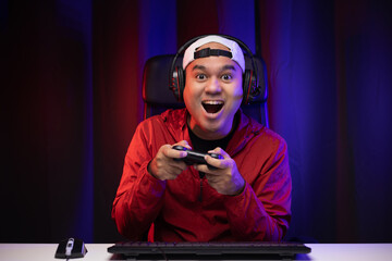 Excited and shocked face of Asian gamer with headphone holding joystick playing video game online sitting on chair at living room. Indian professional gamer streaming on social playing game very fun © Chanakon