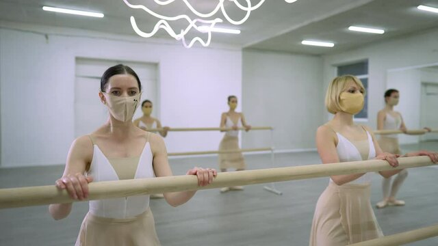 Wide shot ballerinas in coronavirus face masks rehearsing plie and releve movement at barre in dance studio. Young Caucasian women dancing on Covid-19 pandemic lockdown. New normal choreography
