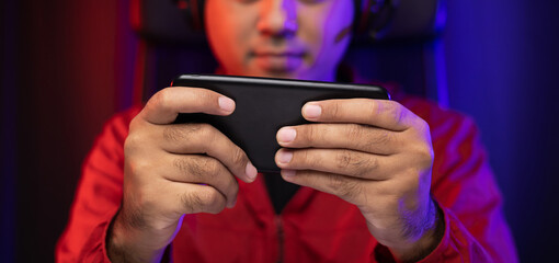 Playing video game on smartphone. Young asian handsome man sitting on chair holding cellphone in living room. Happiness Streamer Indian man wearing headset playing game online in the darkroom. © Chanakon