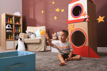 Cute little boy playing on floor near cardboard rocket  at home. Child's room interior