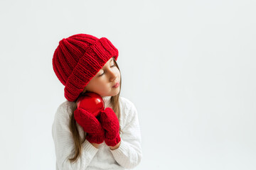 A girl in a red knitted hat and mittens with a Christmas ball. A five-year-old child hugs a ball to himself enjoying the moment. The concept of Christmas and New Year. White background with copy spase