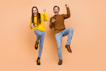 Full length photo of brunette astonished couple hold telephones wear sweaters jeans shoes isolated on beige background