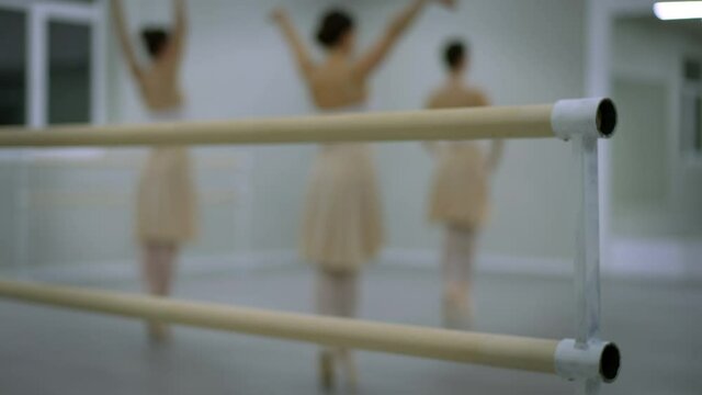 Close-up dancing barre with blurred ballerinas rehearsing eleve at background indoors. Three slim Caucasian agile women dancing classical ballet dance. Rehearsal and talent concept