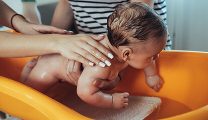Two Anonymous Female Hands Washing Baby Boy at Home. 

Unrecognizable mother having help while giving a happy newborn baby a bath in orange baby bathtub at home.