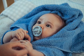 Baby Boy whit Pacifier Wrapped in a Towel After Bath at Home. 

Close up photo of an anonymous mother holding hands of her wet baby lying down on a baby bed.